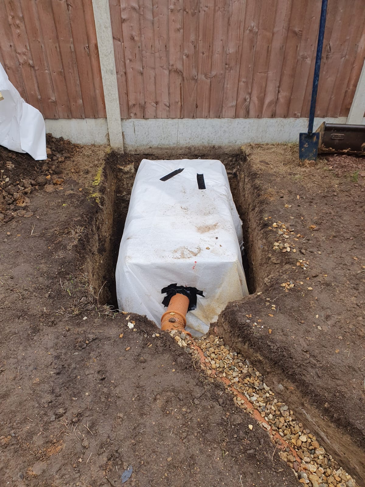 Soakaway crate wrapped in white geotextile membrane sitting in an open trench, with an orange drainage pipe taped into its side and a gravel-filled channel leading to it, beside a wooden fence on concrete posts