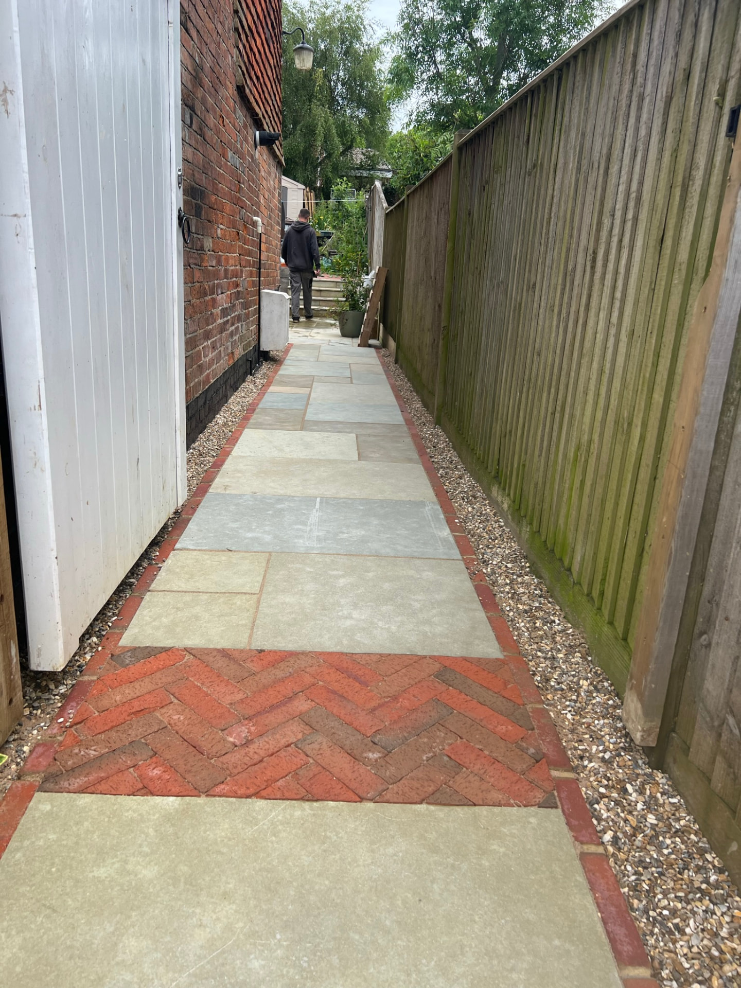 Side access path of large stone slabs with a herringbone red brick section, bordered by pebbles between a white gate and a wooden fence