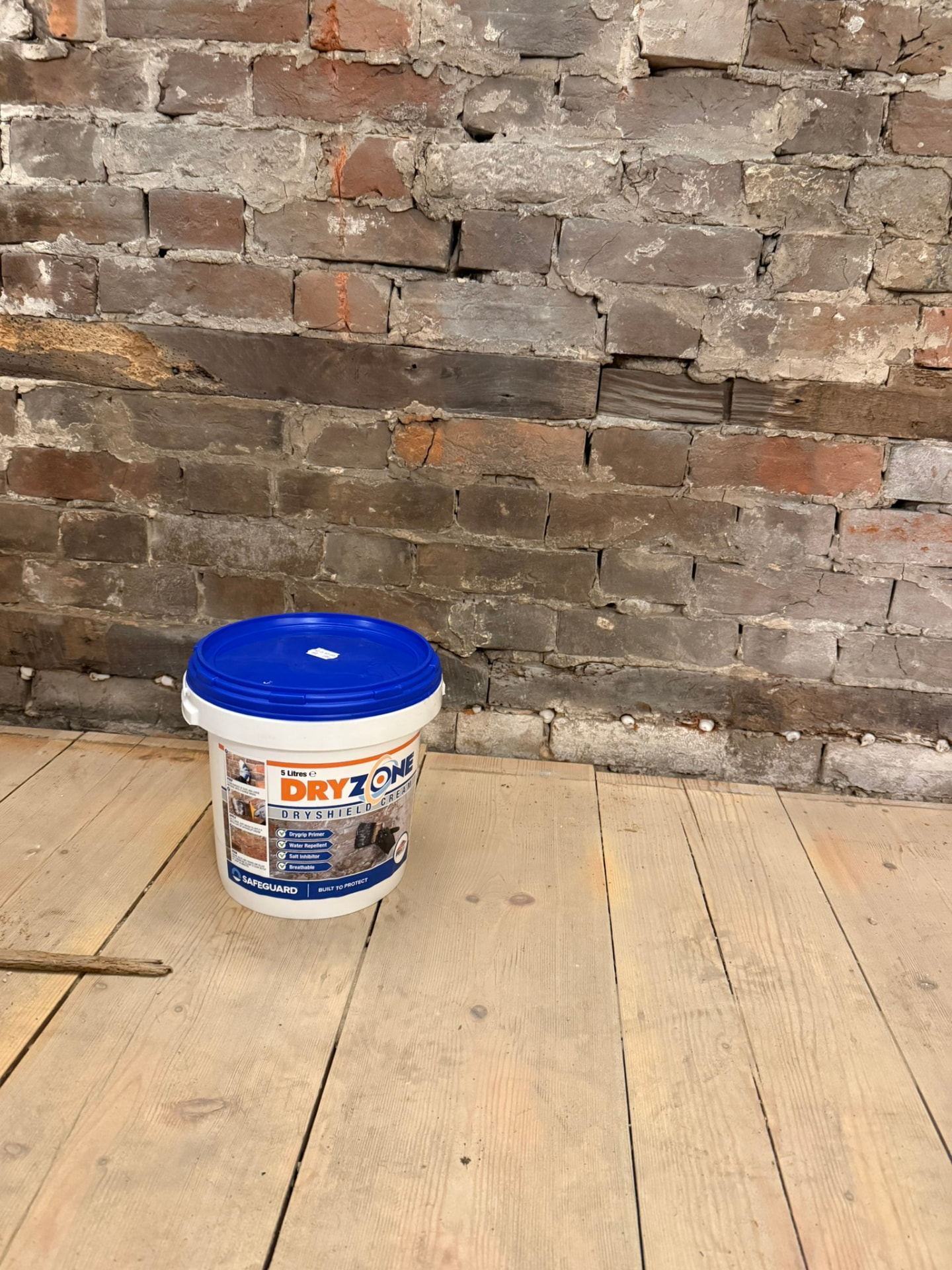 Interior of a period property with a wall stripped back to bare brickwork above bare timber floorboards during damp proofing work, with a tub of damp-proofing cream on the floor