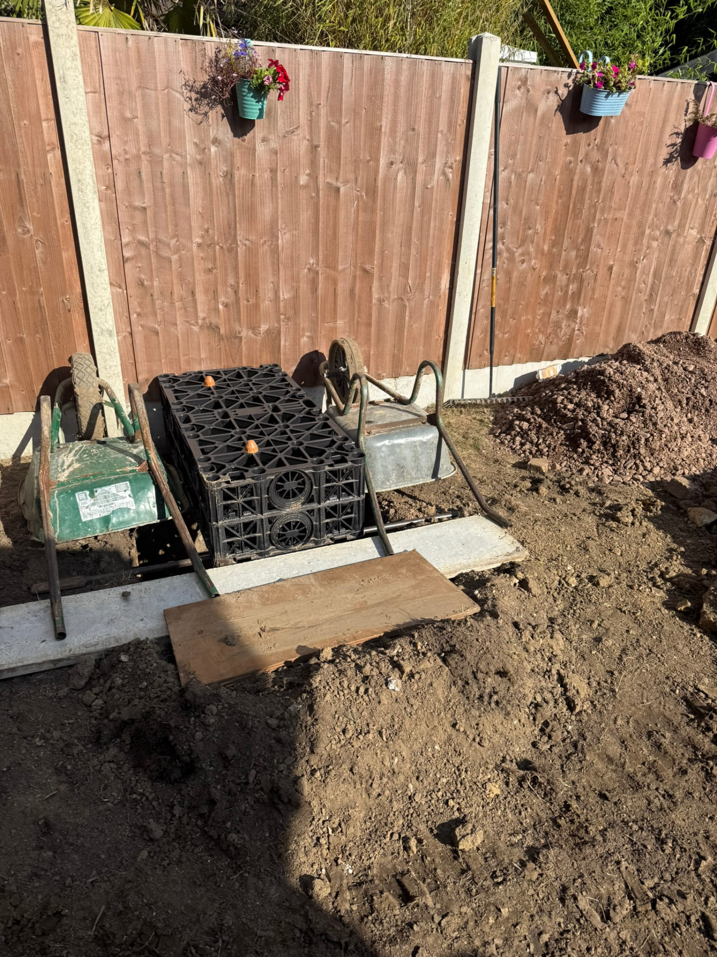 Garden drainage installation in progress with stacked black soakaway crates, wheelbarrows and spoil beside a wooden fence