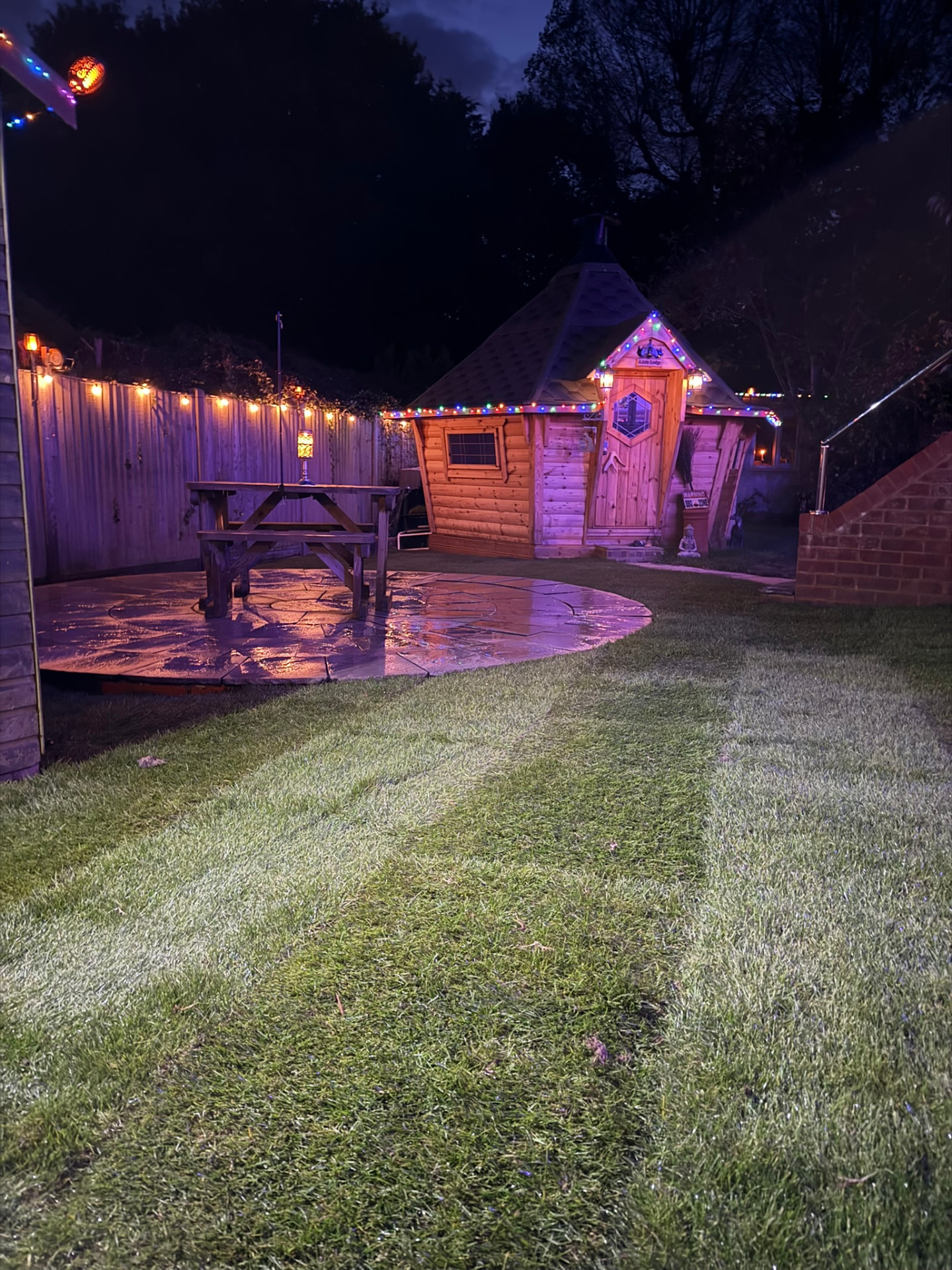 Stone patio with a wooden picnic table beside a garden room decorated with string lights at night